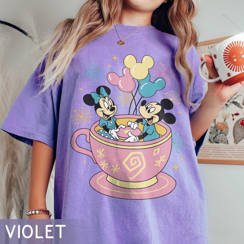 May include: A violet-coloured t-shirt with a playful illustration of Mickey and Minnie Mouse in a teacup, accompanied by balloons and fireworks. The shirt has a loose fit, and the word "VIOLET" is printed at the bottom.