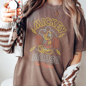 Puede incluir: Camiseta marr&oacute;n con un gr&aacute;fico de Mickey Mouse, con las palabras "Mickey Mouse" en amarillo y azul. El dise&ntilde;o incluye los n&uacute;meros "28" en rojo y la frase "The Original" en blanco.