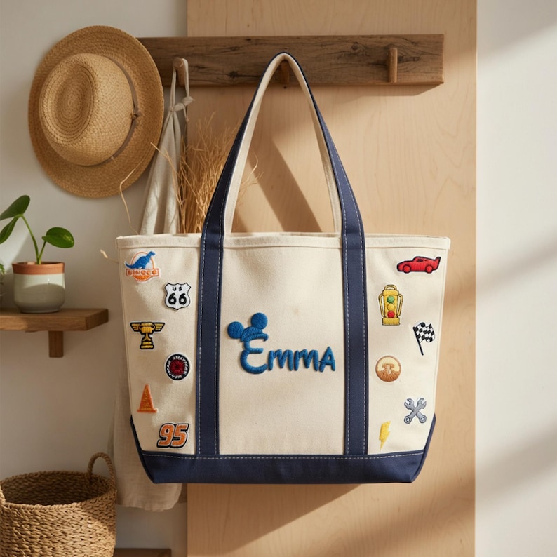 May include: A canvas tote bag with a cream-colored body and navy blue trim. The bag features embroidered patches of cars, a racing flag, and the name "Emma" in blue. A straw hat hangs on a wooden wall in the background.