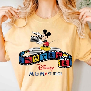 Pu&ograve; includere: T-shirt gialla con Topolino che tiene una clapperboard con la scritta "ACTION". Il design include una bobina di film e una striscia di film colorata con il marchio Disney MGM Studios.