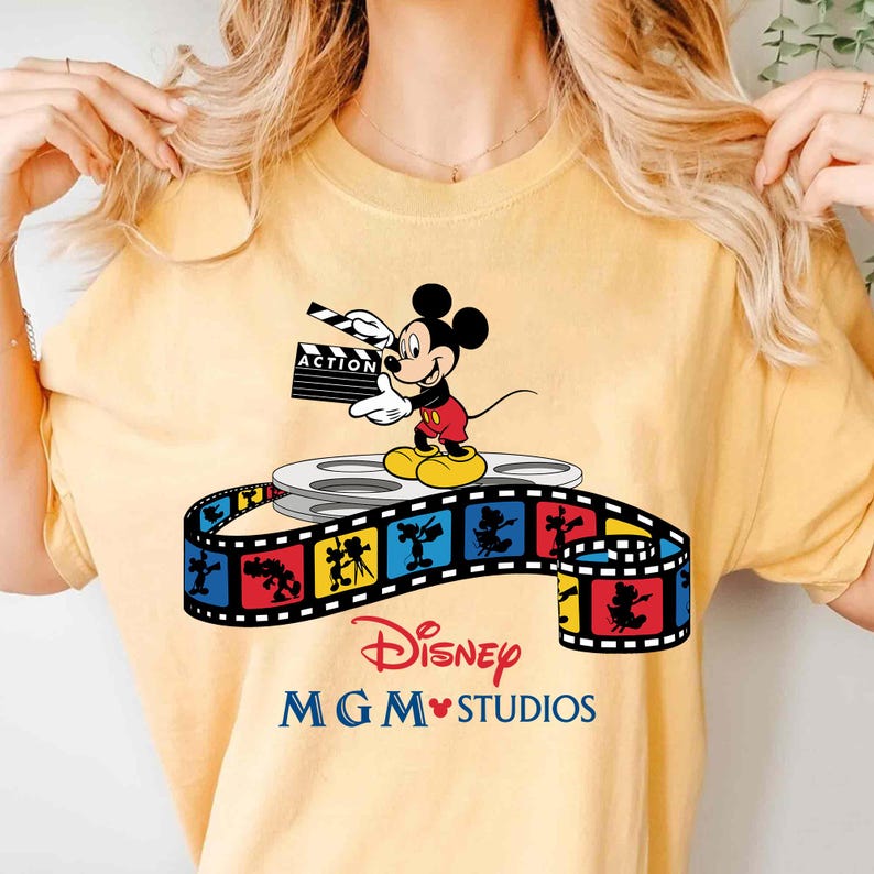 Pu&ograve; includere: T-shirt gialla con Topolino che tiene una clapperboard con la scritta "ACTION". Il design include una bobina di film e una striscia di film colorata con il marchio Disney MGM Studios.