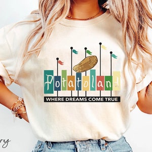 Könnte beinhalten: Elfenbeinfarbenes T-Shirt mit dem Wort "Potatoland" in bunten Blockbuchstaben, einer Cartoon-Kartoffel und Flaggen. Der Text "WHERE DREAMS COME TRUE" steht darunter. Das Shirt hat eine hellcremefarbene Farbe.
