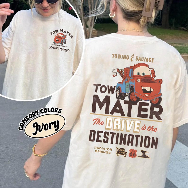 May include: Ivory-colored t-shirt featuring a graphic of Tow Mater from the movie Cars. The design includes the text "TOW MATER," "The DRIVE is the DESTINATION," and "Radiator Springs." The shirt is a Comfort Colors brand.