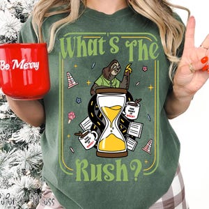 Puede incluir: Camiseta verde con un dise&ntilde;o caprichoso que presenta un perezoso, un reloj de arena y el texto "What's The Rush?". El dise&ntilde;o incluye tazas de caf&eacute; y conos de tr&aacute;fico. En la imagen se sostiene una taza roja con "Be Merry".