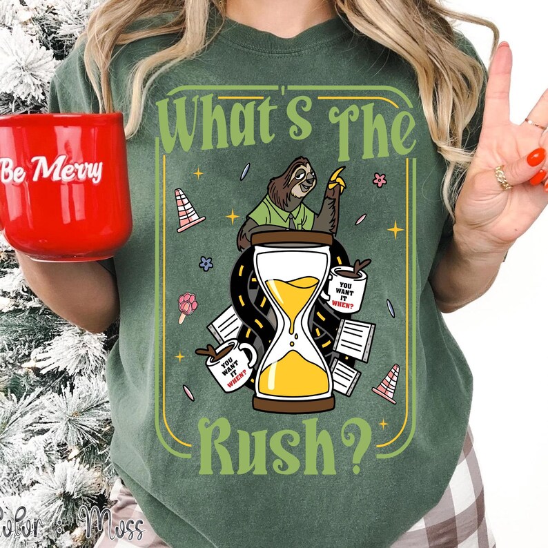 Puede incluir: Camiseta verde con un dise&ntilde;o caprichoso que presenta un perezoso, un reloj de arena y el texto "What's The Rush?". El dise&ntilde;o incluye tazas de caf&eacute; y conos de tr&aacute;fico. En la imagen se sostiene una taza roja con "Be Merry".