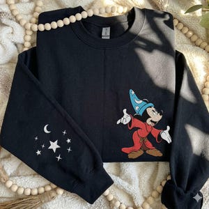 Puede incluir: Una sudadera negra de cuello redondo con un bordado de un rat&oacute;n de dibujos animados que lleva un sombrero de mago azul y una t&uacute;nica roja. La manga izquierda est&aacute; decorada con un patr&oacute;n bordado de una luna creciente blanca y estrellas.