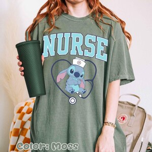 May include: Moss green t-shirt with the word "NURSE" in light blue block letters. A cartoon character wearing a nurse's hat is inside a heart-shaped stethoscope. A dark green studded tumbler is held in the left hand.