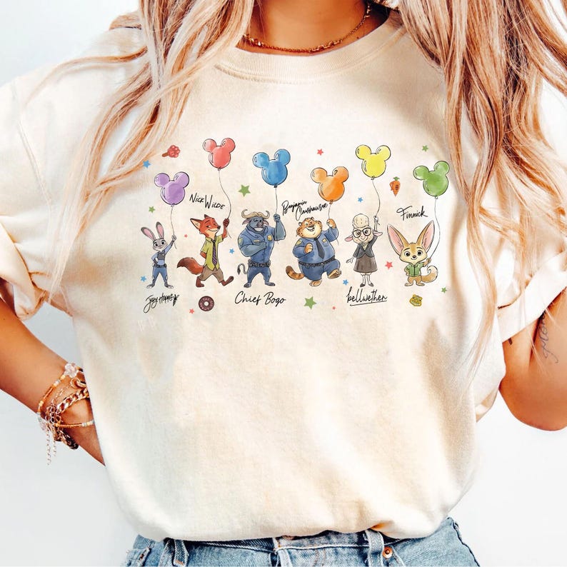 Op de afbeelding: Een beige sweatshirt met cartoonfiguren uit Zootopia die Mickey Mouse-vormige ballonnen vasthouden. De personages zijn Judy Hopps, Nick Wilde, Chief Bogo, Benjamin Clawhauser, Bellwether en Finnick. Tekst bevat de namen van de personages.