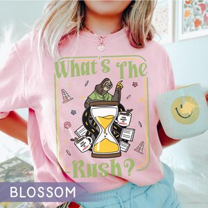 Puede incluir: Una camiseta rosa claro con el texto "What's The Rush?" y un gr&aacute;fico de un perezoso, un reloj de arena y tazas de caf&eacute;. La camiseta tambi&eacute;n tiene la palabra "BLOSSOM" en la parte inferior. En la imagen se sostiene una taza con una carita sonriente.