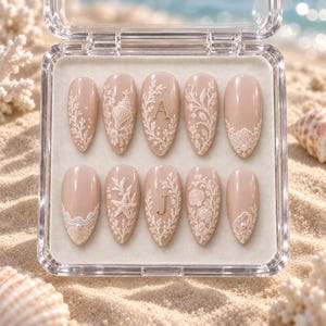May include: A set of ten almond-shaped press-on nails in a nude beige tone, displayed in a clear plastic case. The nails feature intricate white raised relief designs of seashells, starfish, coral, and lace patterns, with gold letters A and J.
