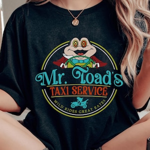 May include: Black t-shirt with a cartoon image of Mr. Toad driving a car. The text reads "Mr. Toad's Taxi Service" with the words "Wild Rides Great Rates" around the circle.