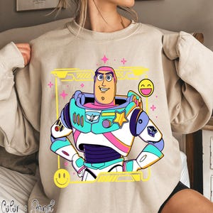 May include: Beige sweatshirt featuring a colorful cartoon graphic of Buzz Lightyear. The graphic includes a yellow frame, pink accents, and smiley face emojis. The design is a vibrant and playful depiction of the character.