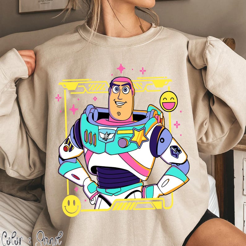 May include: Beige sweatshirt featuring a colorful cartoon graphic of Buzz Lightyear. The graphic includes a yellow frame, pink accents, and smiley face emojis. The design is a vibrant and playful depiction of the character.