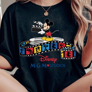 Pu&ograve; includere: T-shirt nero con Topolino che tiene una clapperboard con la scritta "ACTION". La maglietta presenta un design a striscia di film colorato con i loghi Disney e MGM Studios.