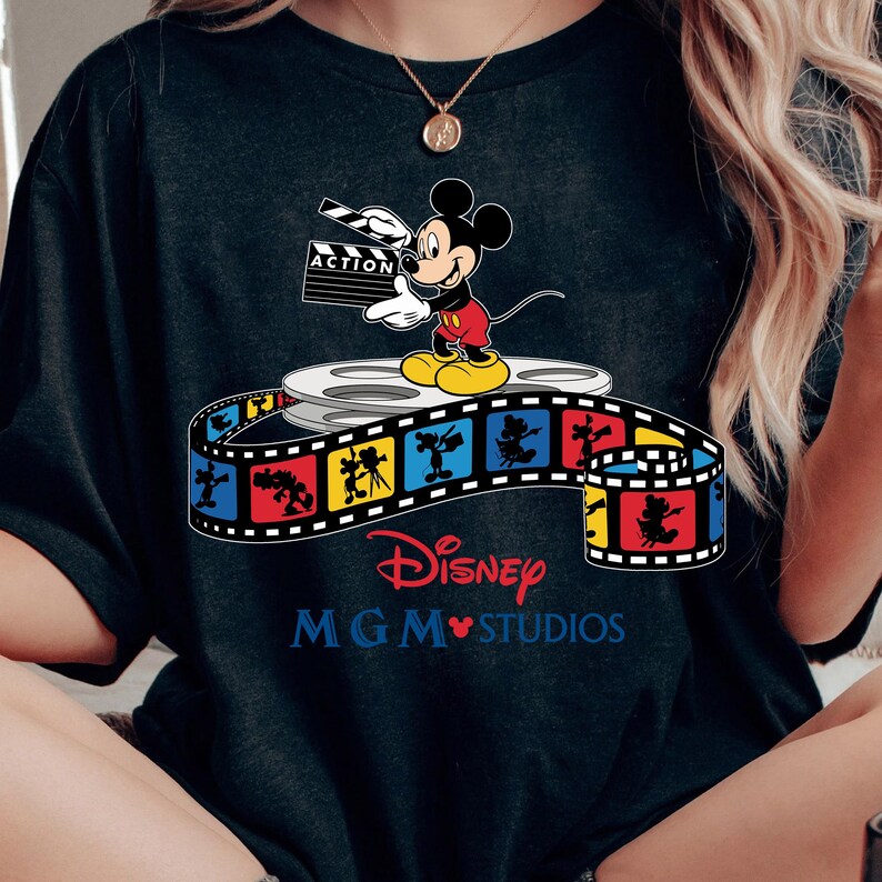 Pu&ograve; includere: T-shirt nero con Topolino che tiene una clapperboard con la scritta "ACTION". La maglietta presenta un design a striscia di film colorato con i loghi Disney e MGM Studios.