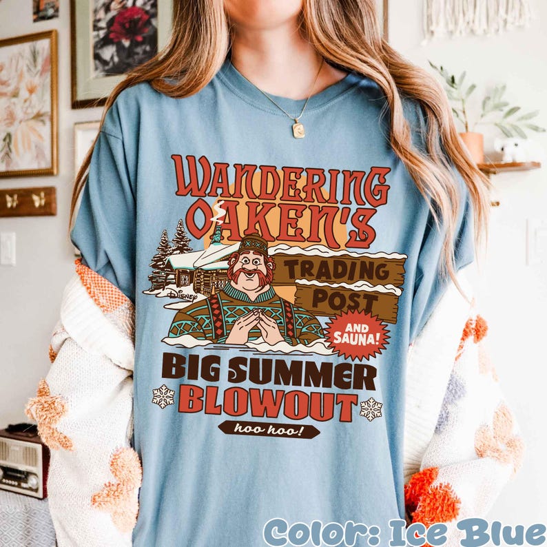 Op de afbeelding: IJsblauw T-shirt met een grafisch ontwerp. Het ontwerp bevat de tekst "Wandering Oaken's Trading Post" en een stripfiguur. De woorden "Big Summer Blowout" en "hoo hoo!" zijn ook aanwezig.
