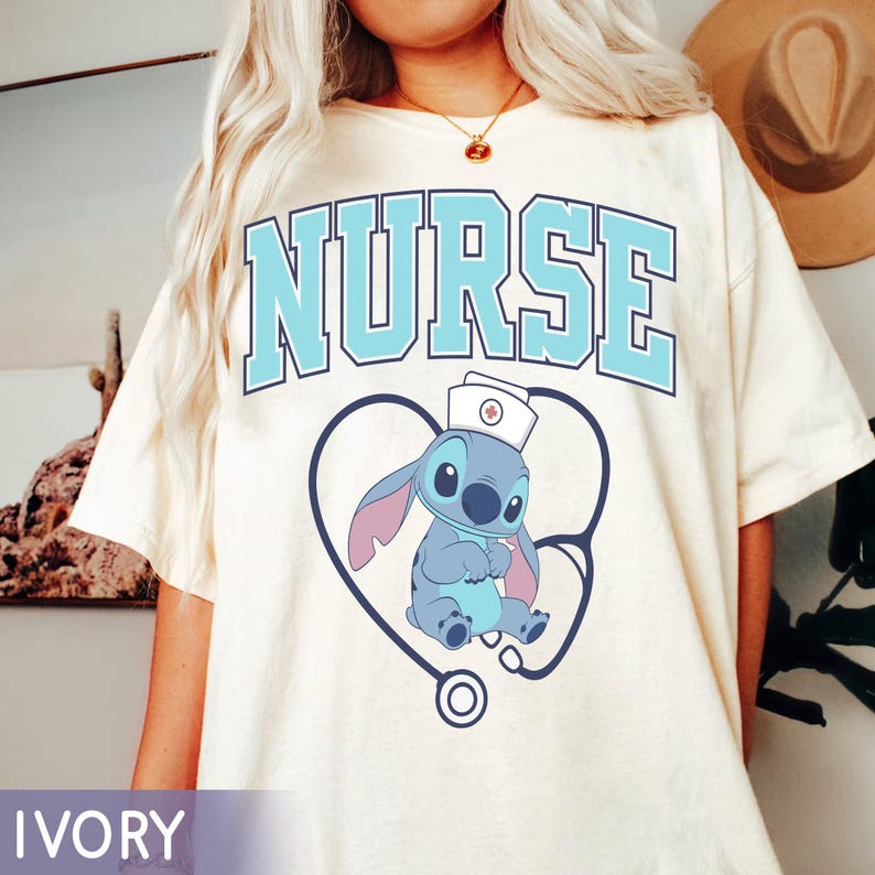 May include: Ivory-colored t-shirt with the word "NURSE" in light blue block letters. Below the text is a blue cartoon character wearing a nurse's cap, inside a heart-shaped stethoscope.