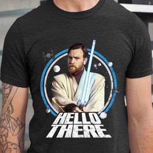 May include: A dark gray t-shirt with a blue and white circle graphic featuring a character from Star Wars holding a lightsaber. The text "HELLO THERE" is printed in white below the graphic.