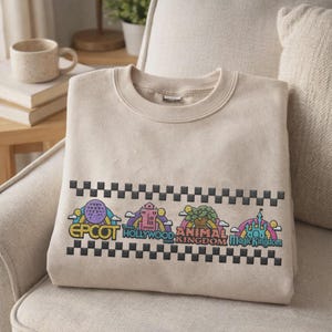 Puede incluir: Sudadera beige con cuello redondo, con gr&aacute;ficos bordados coloridos de iconos de parques tem&aacute;ticos. El dise&ntilde;o incluye las palabras "EPCOT", "HOLLYWOOD", "ANIMAL KINGDOM" y "Magic Kingdom" sobre un patr&oacute;n de cuadros blancos y negros.