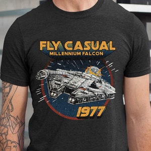Puede incluir: Una camiseta gris oscuro con un gráfico de estilo vintage del Millennium Falcon, la nave espacial de Star Wars. El gráfico incluye el texto "Fly Casual" y "Millennium Falcon" y el año "1977".