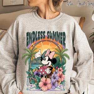 May include: Sport grey sweatshirt featuring a colorful graphic of Minnie Mouse in a pink polka dot dress, with the words "ENDLESS SUMMER" above her. The design includes palm trees, flowers, and a sunset scene. The sweatshirt is a comfortable, casual style.