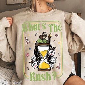 Puede incluir: Sudadera color arena con un gr&aacute;fico que presenta un perezoso se&ntilde;alando hacia arriba, un reloj de arena y el texto "What's The Rush?". El dise&ntilde;o incluye tazas de caf&eacute; y elementos de carretera.