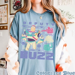 May include: Ice blue t-shirt featuring a graphic of Buzz Lightyear with the text "DEPUTY BUZZ". The design includes colorful illustrations of Buzz and other characters. The shirt is a light blue color.