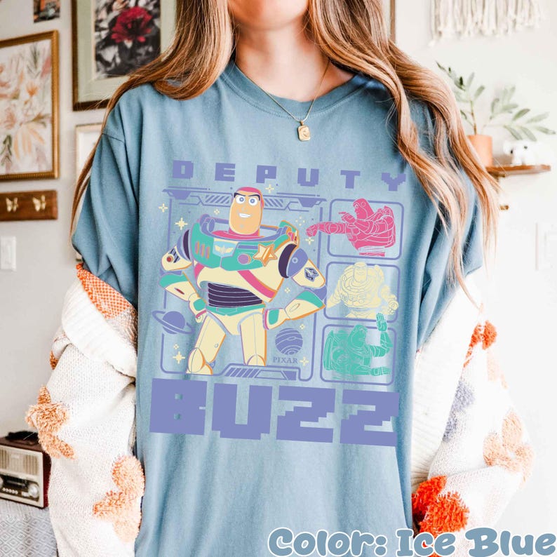 May include: Ice blue t-shirt featuring a graphic of Buzz Lightyear with the text "DEPUTY BUZZ". The design includes colorful illustrations of Buzz and other characters. The shirt is a light blue color.