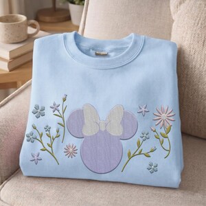 Pu&ograve; includere: Una felpa girocollo azzurra con una silhouette ricamata di una testa di topo viola con un fiocco bianco al centro. Il disegno &egrave; circondato da vari steli floreali ricamati e piccoli fiori nelle tonalit&agrave; del blu e del rosa.