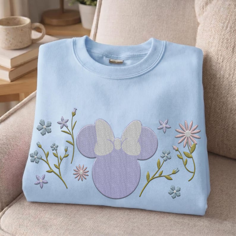 Pu&ograve; includere: Una felpa girocollo azzurra con una silhouette ricamata di una testa di topo viola con un fiocco bianco al centro. Il disegno &egrave; circondato da vari steli floreali ricamati e piccoli fiori nelle tonalit&agrave; del blu e del rosa.