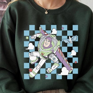 May include: Green sweatshirt with a cartoon image of Buzz Lightyear from Toy Story flying in a spaceship. The image is on a checkered background with clouds and stars.