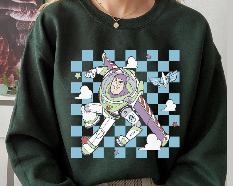 May include: Green sweatshirt with a cartoon image of Buzz Lightyear from Toy Story flying in a spaceship. The image is on a checkered background with clouds and stars.