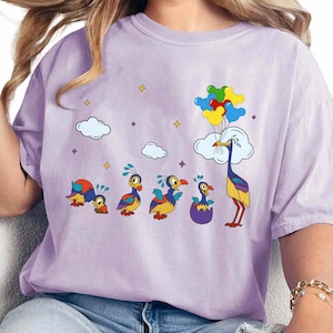 May include: Purple t-shirt with a cartoon image of a family of birds. The birds are blue, yellow, and orange. The father bird is holding a bunch of colorful balloons. The shirt has a light purple background with white clouds and stars.