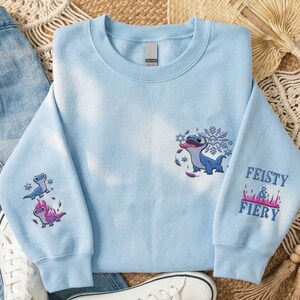 May include: A light blue crewneck sweatshirt featuring embroidered designs of a blue lizard character. The chest shows the character with snowflakes, while the sleeves display the character with flames and the text Feisty & Fiery in blue.