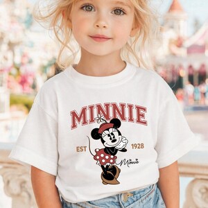 May include: White t-shirt featuring a Minnie Mouse graphic. The graphic includes the name "MINNIE" in maroon block letters, the text "EST 1928", and an illustration of Minnie Mouse in a red polka-dot dress. The shirt is worn by a young child.