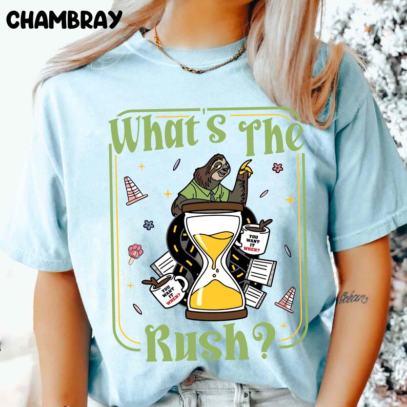 Puede incluir: Camiseta azul claro con un gr&aacute;fico que presenta un perezoso, un reloj de arena y el texto "What's The Rush?". El dise&ntilde;o incluye tazas de caf&eacute; y elementos de carretera. La camiseta es de material suave.
