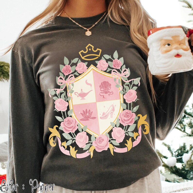 Puede incluir: Una camiseta de manga larga gris oscuro con un dise&ntilde;o de escudo con rosas rosas, lazos y una corona. El escudo tiene un patr&oacute;n a cuadros rosa y blanco con ilustraciones de una l&aacute;mpara, una concha, una flor de loto y un tac&oacute;n alto. El texto "Color: Pepper" est&aacute; en la parte inferior.