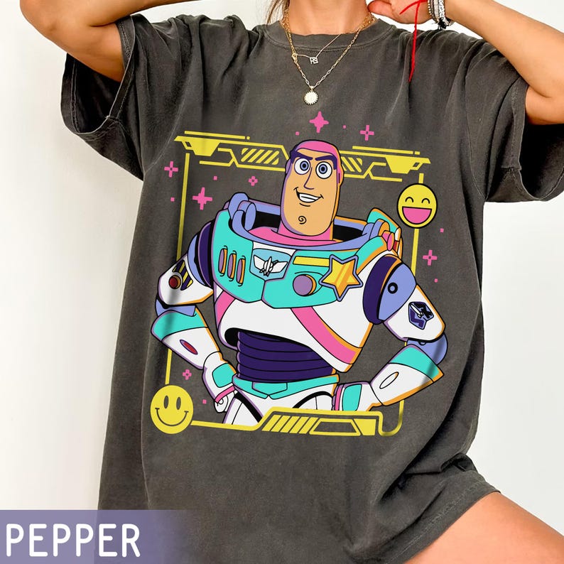 May include: Dark gray t-shirt featuring a colorful graphic of Buzz Lightyear from Toy Story. The design includes a yellow frame, stars, and smiley faces. The word "PEPPER" is printed at the bottom.