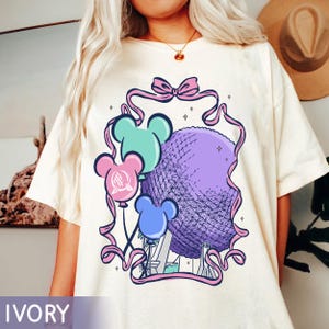 May include: Ivory-colored t-shirt featuring a graphic of a purple sphere, reminiscent of a geodesic dome, with Mickey Mouse ear balloons in teal, pink, and blue. The design is framed by a pink ribbon with a bow at the top. The word "IVORY" is at the bottom.