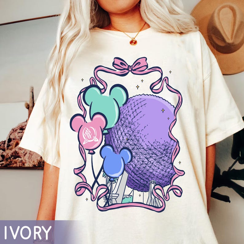 May include: Ivory-colored t-shirt featuring a graphic of a purple sphere, reminiscent of a geodesic dome, with Mickey Mouse ear balloons in teal, pink, and blue. The design is framed by a pink ribbon with a bow at the top. The word "IVORY" is at the bottom.