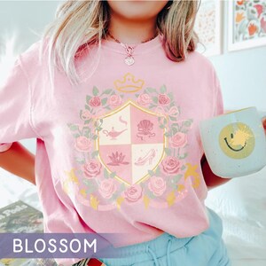 Puede incluir: Camiseta rosa claro con un dise&ntilde;o de escudo con una corona, rosas e iconos de cuentos de hadas. La camiseta tiene cuello redondo y mangas cortas. Una taza azul claro con una carita sonriente amarilla se sostiene en la mano. La palabra "BLOSSOM" est&aacute; en la parte inferior.
