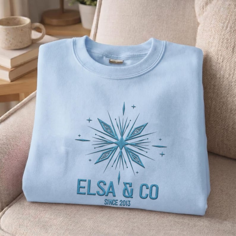 May include: A light blue crewneck sweatshirt featuring an embroidered dark blue snowflake design centered on the chest. Below the snowflake, the text reads ELSA & CO in a bold font, with the smaller text SINCE 2013 positioned underneath.