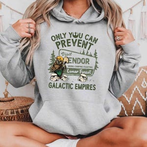 Puede incluir: Sudadera gris con capucha con un estampado gráfico verde que dice "Only you can prevent Galactic Empires" y presenta un personaje de dibujos animados con el texto "Visit Endor Hiking-Camping-Feasting Lunar Forest".