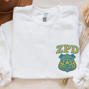 May include: A white crewneck sweatshirt featuring an embroidered ZPD Zootopia Police badge logo on the lower right side. The badge is designed in yellow and blue thread, with the text ZPD positioned above the shield-shaped police emblem.