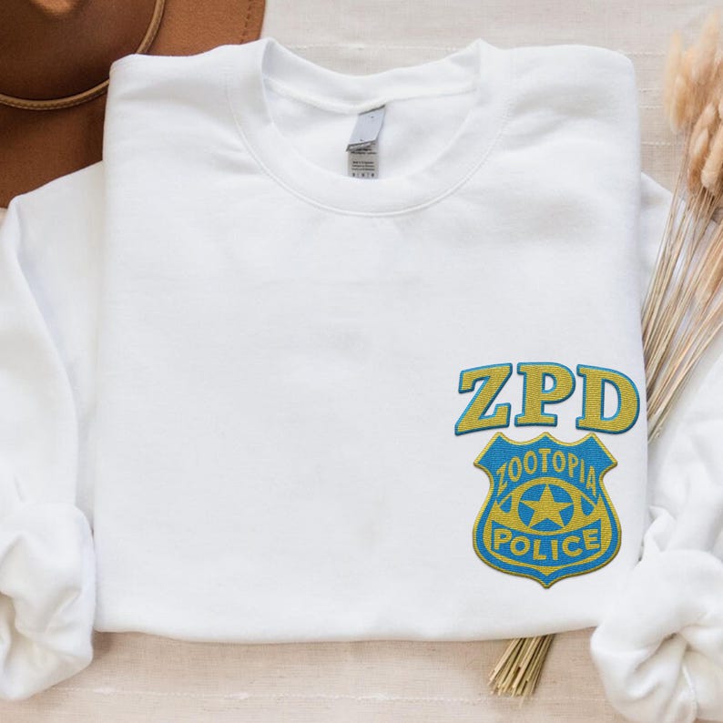May include: A white crewneck sweatshirt featuring an embroidered ZPD Zootopia Police badge logo on the lower right side. The badge is designed in yellow and blue thread, with the text ZPD positioned above the shield-shaped police emblem.