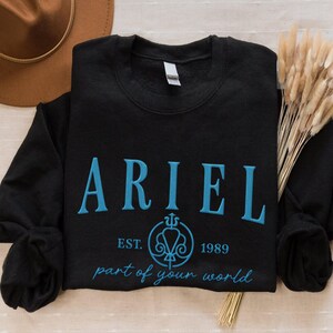 May include: Black crewneck sweatshirt with the name "ARIEL" embroidered in light blue, along with the text "EST. 1989" and "part of your world". The sweatshirt is folded, with a brown hat and dried plants in the background.