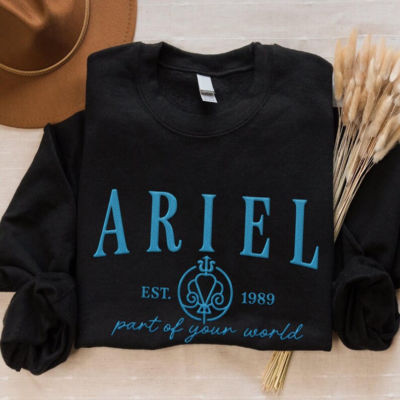 May include: Black crewneck sweatshirt with the name "ARIEL" embroidered in light blue, along with the text "EST. 1989" and "part of your world". The sweatshirt is folded, with a brown hat and dried plants in the background.
