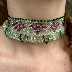 Op de afbeelding: Een kralen choker met een groen en wit patroon en paarse accenten. De ketting heeft een franje van groene kralen.