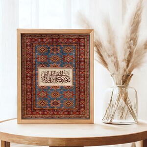 May include: A framed print featuring a detailed rug design in shades of blue, red, and gold. The print includes Arabic calligraphy in a central rectangular panel. The wooden frame sits on a light-colored wooden table.