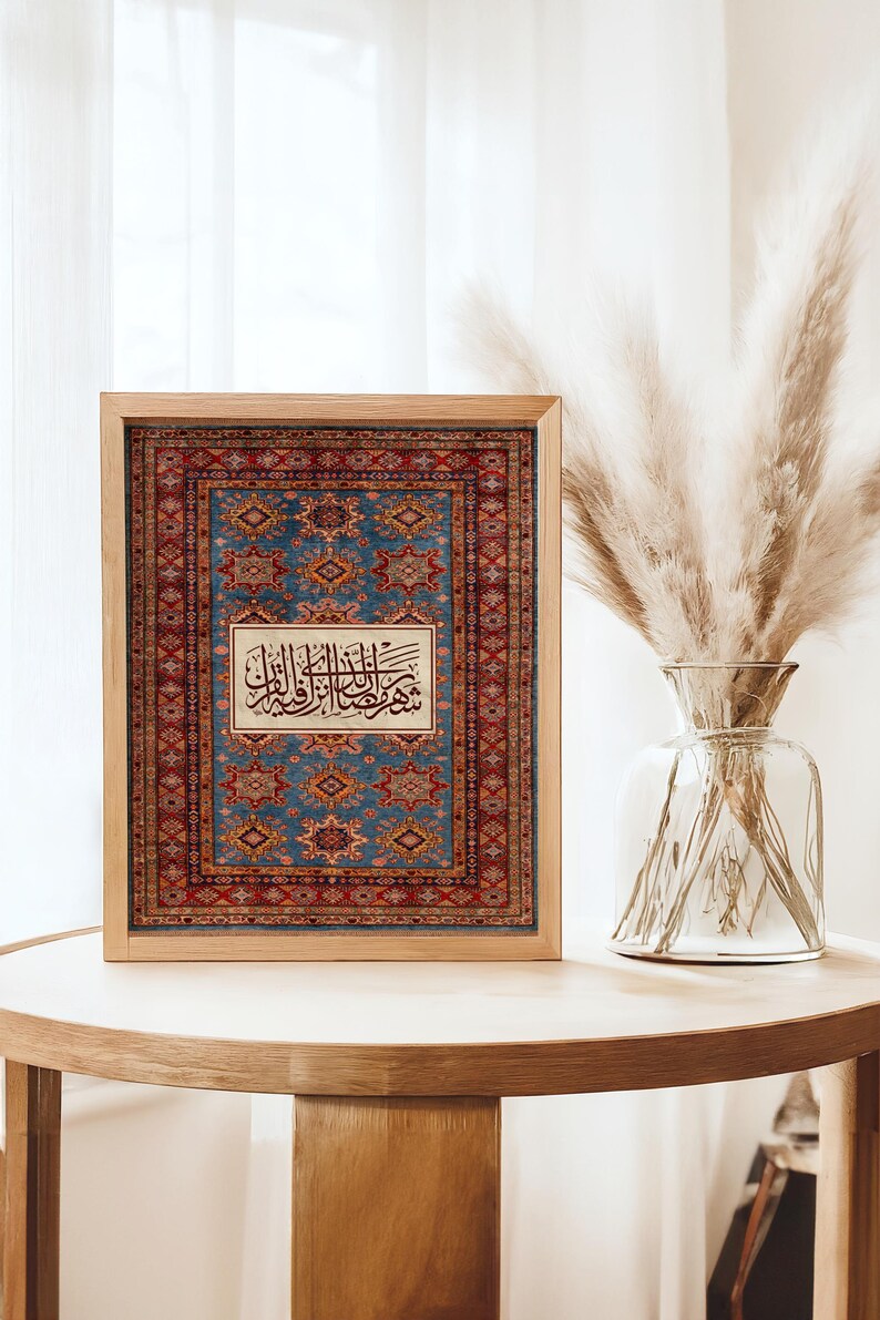 May include: A framed print featuring a detailed rug design in shades of blue, red, and gold. The print includes Arabic calligraphy in a central rectangular panel. The wooden frame sits on a light-colored wooden table.